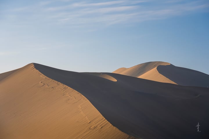 A solitary figure stands on a sandy hill in a vast desert landscape, with a clear blue sky above.