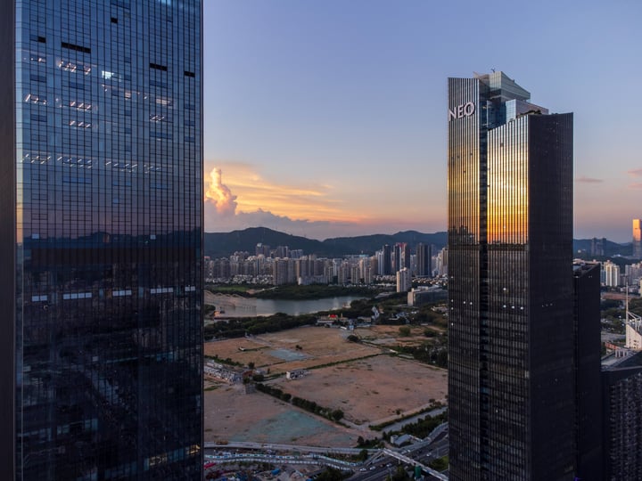 A cityscape at sunset, with three tall modern skyscrapers reflecting the setting sun, a river, and mountains in the background.