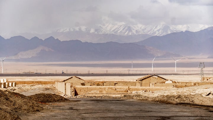 A desert landscape with snow-capped mountains in the background, a dirt road, and a few buildings with straw roofs, with wind turbines in the distance.
