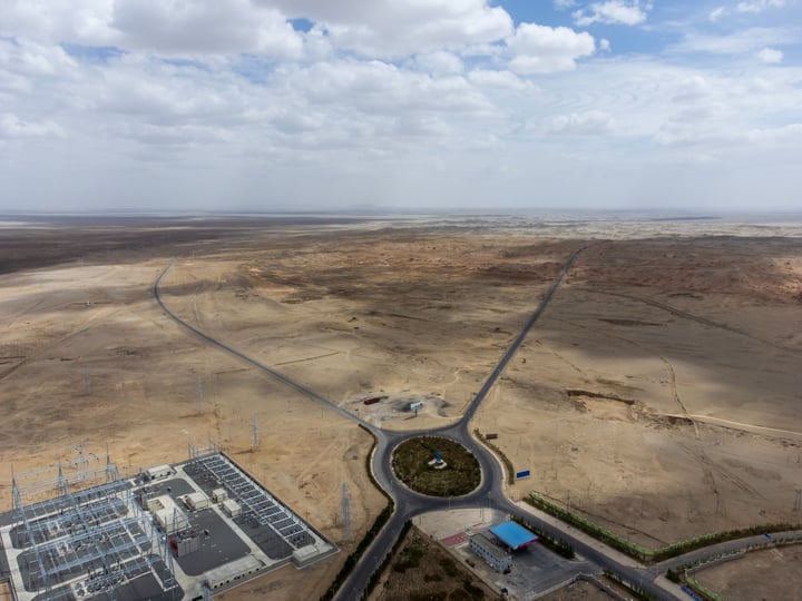 An aerial view of a large power plant with wind turbines, surrounded by a road and a blue building, set against a vast, barren landscape with a clear blue sky.
