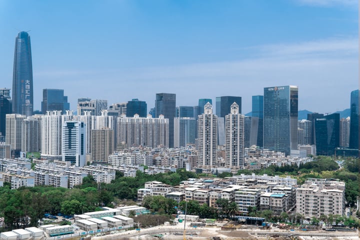 A cityscape with tall buildings, including a prominent skyscraper, is captured from a high vantage point. The sky is clear blue, and the buildings are predominantly gray and white. Trees are scattered throughout the foreground, adding a touch of nature to the urban landscape.