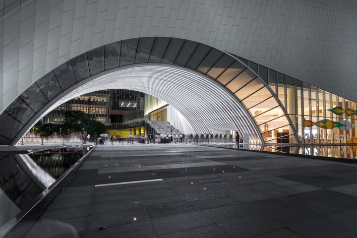 A modern, white, curved building with a glass facade stands in a cityscape, its curved roof and reflective glass windows reflecting the surroundings. A walkway leads to the building, with a body of water nearby. People walk on the sidewalk, and a yellow building is visible in the background.