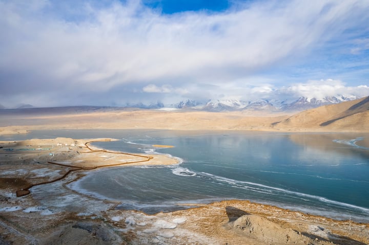A serene landscape features a deep blue lake reflecting the sky, with snow-capped mountains in the distance. The sky is filled with white clouds, and the landscape is dominated by brown and beige tones.