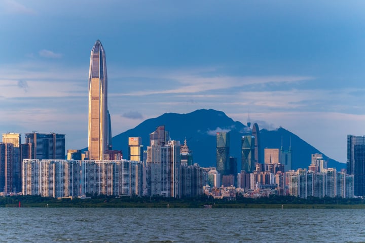A panoramic view of a bustling cityscape with tall buildings, a prominent skyscraper, and a mountain in the background. The sky is a clear blue with scattered clouds.