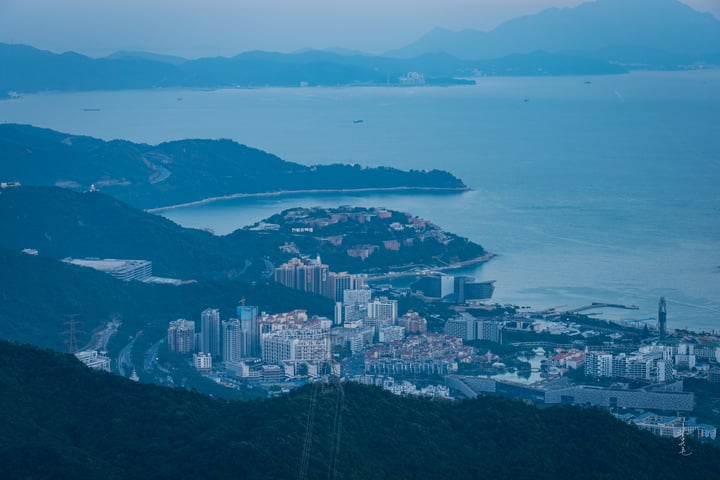 A panoramic view of a city nestled on a hillside, surrounded by mountains and a large body of water, with a few boats on the water and a few buildings in the cityscape.