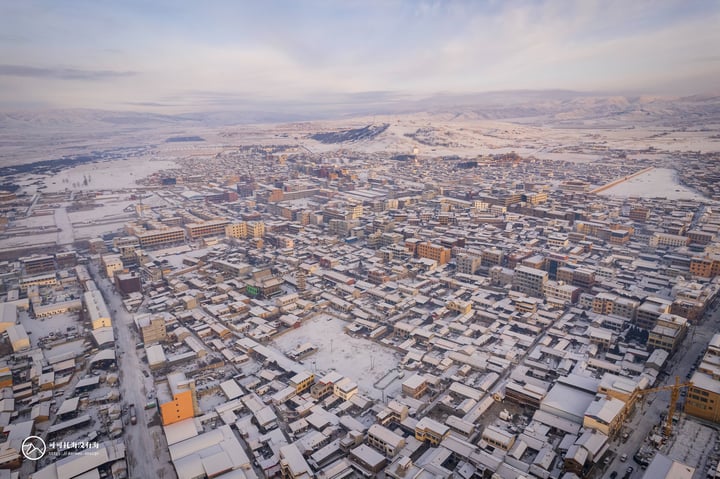 An aerial view of a snowy city with densely packed buildings, roads, and a mountain range in the background.