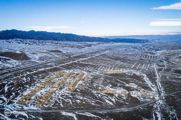 An aerial view of a snowy landscape with a mountain range in the background, a road winding through the terrain, and a clear blue sky above.