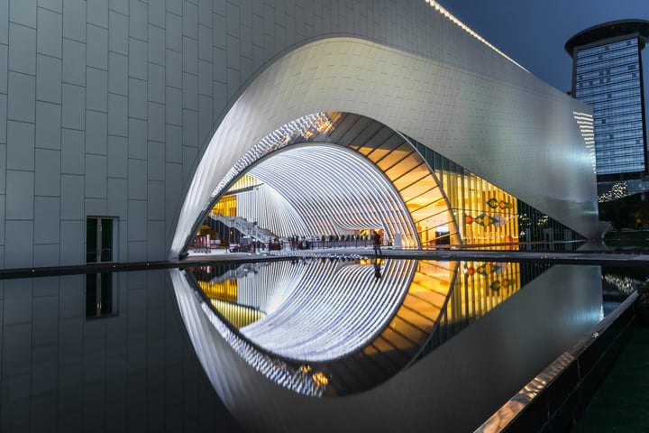 A modern building with a curved white metal archway and reflective pool in front, illuminated by white lights, is surrounded by a dark gray building and a solitary figure walking on the sidewalk.