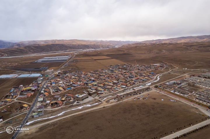 An aerial view of a rural area with a mix of buildings, roads, and fields, surrounded by mountains and clouds.