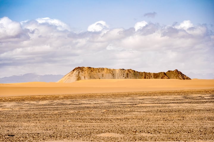 A vast desert landscape with a large sand mound in the foreground, a clear blue sky with white clouds, and a distant mountain range.