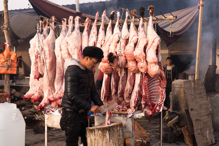 A man in a black leather jacket is preparing meat at a market stall, with a table of raw meat and a bucket nearby. The stall is set up in a market setting with a tent and other stalls in the background.