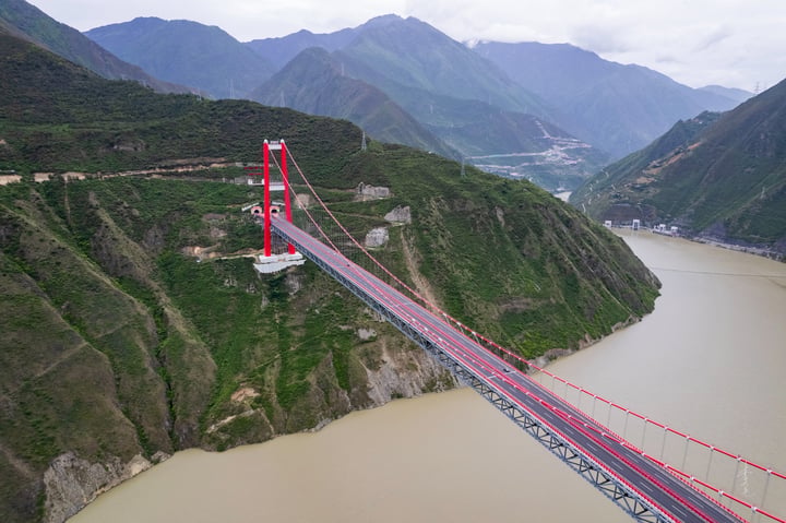 A red and white suspension bridge spans a river, with a white cable connecting the two sides. The bridge is surrounded by mountains and green hills, and the sky is cloudy.