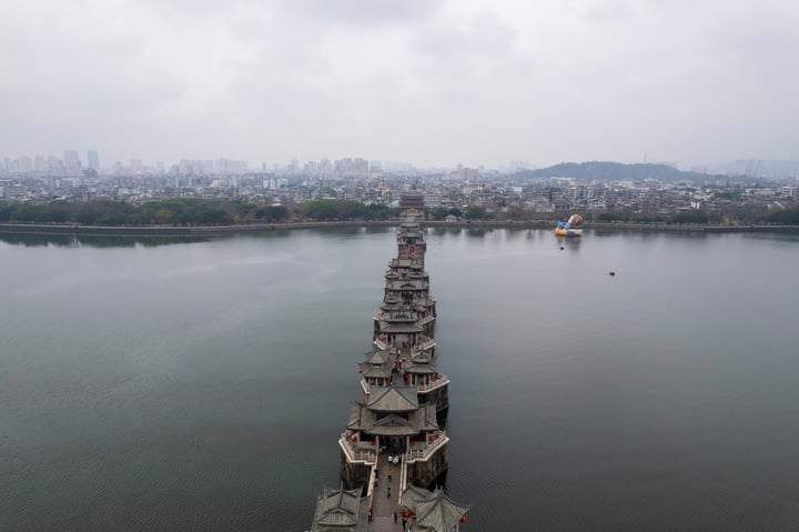 一座传统风格的桥梁延伸至水面远方，背景是多云的天空和远处的城市建筑景观。