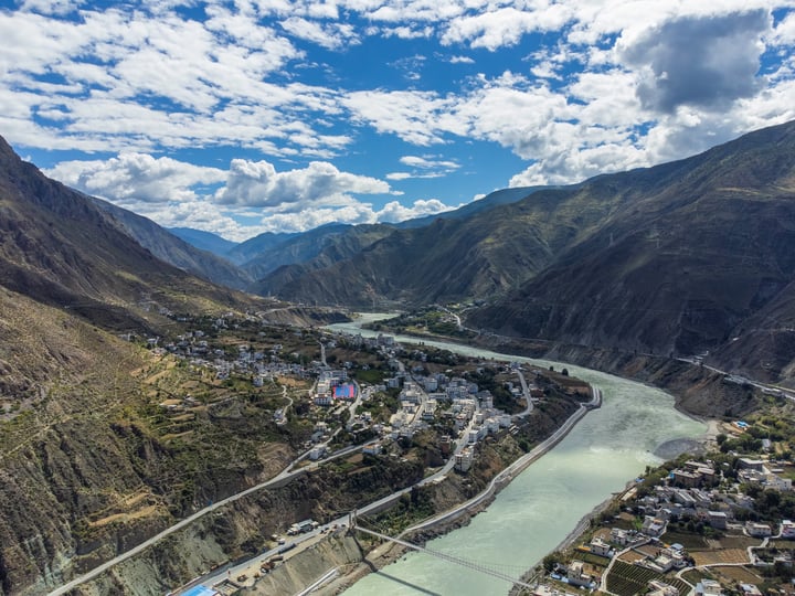 An aerial view of a picturesque valley with a river, surrounded by mountains and buildings, with a bridge connecting the two sides.