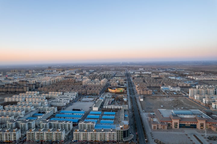 An aerial view of a densely populated city with a mix of modern and older buildings, surrounded by a vast expanse of open space and a road. The sky is a mix of blue and orange hues, indicating either sunrise or sunset.