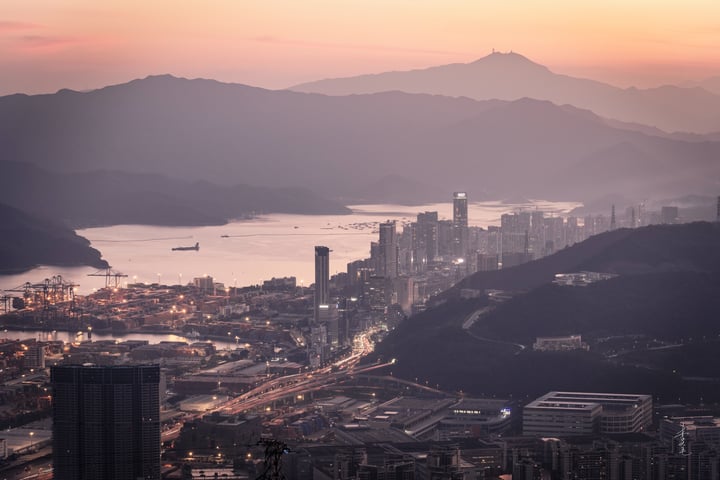 A cityscape at dusk, with tall buildings and a large body of water, a mountain range in the background, and a large ship on the water.