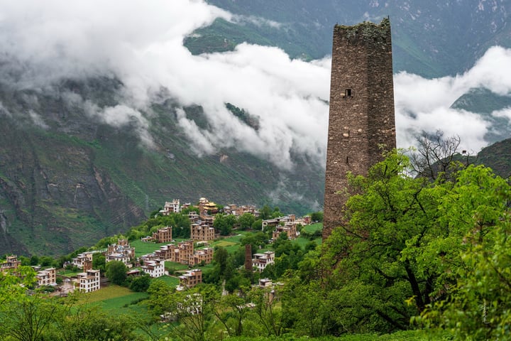 A medieval stone tower stands tall amidst a verdant valley, surrounded by lush greenery and a mountain range. The tower, with its narrow windows and pointed top, is the central focus of the image, while the valley below is dotted with houses and trees. The sky above is filled with clouds, adding a dramatic touch to the scene.