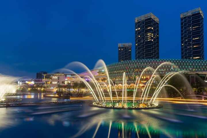 A large fountain with three tiers is illuminated by lights, creating a mesmerizing display of water sprays. The fountain is surrounded by a circular pool, and the background features tall buildings with reflective surfaces. The scene is set against a dark blue sky, emphasizing the fountain's vibrant colors.