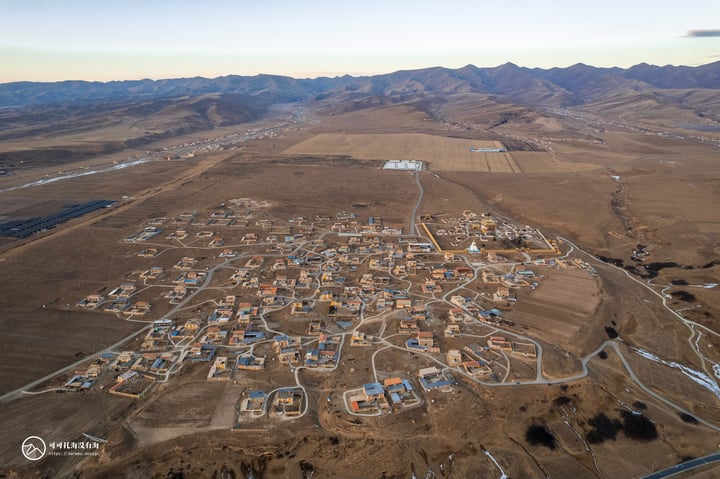 An aerial view of a small town nestled in a desert landscape, surrounded by mountains and a clear blue sky. The town is densely packed with buildings, roads, and fields, and is surrounded by a vast expanse of desert.