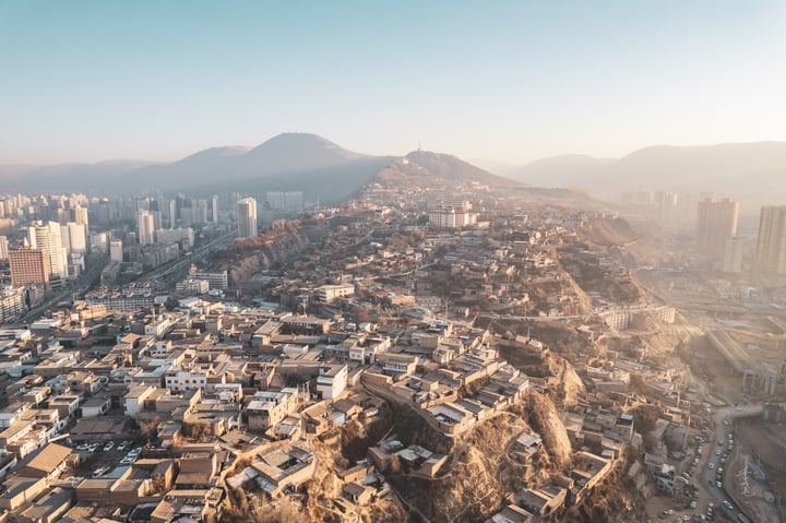 An aerial view of a city nestled on a hillside, surrounded by mountains and a mountain range, with a large mountain in the background. The city is densely populated with white and gray buildings, and the sky is a light blue.