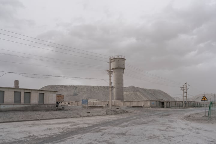 A large white cement tower stands tall in a barren, dusty landscape, with a small white building and a yellow warning sign nearby. A road runs through the scene, with power lines and a utility pole in the distance.