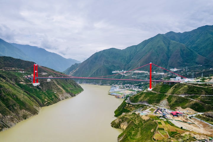 The image captures the iconic Xishan Bridge, a red suspension bridge spanning the Yangtze River in China. The bridge, with its red cables and towers, spans the river, connecting the city of Xishan to the mountains in the background. The river, a brownish-green, flows beneath the bridge, and the mountains, covered in green vegetation, form a majestic backdrop. The sky is cloudy, casting a soft light over the scene. The image is taken from a high vantage point, offering a comprehensive view of the bridge and its surroundings.
