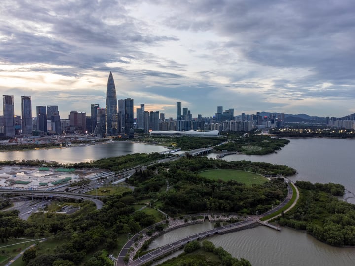 An aerial view of a city skyline with tall buildings, a large body of water, and a bridge, surrounded by greenery and trees.