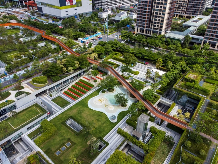 An aerial view of a city park with a winding red train track, surrounded by lush greenery and a white fountain, with tall buildings in the background.