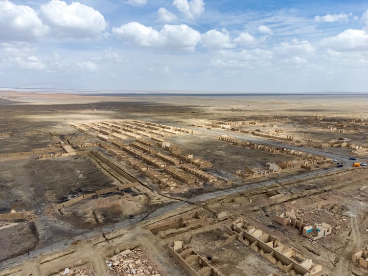 An aerial view of a desert landscape with a large expanse of dirt and a few scattered buildings, with a blue sky and clouds above.