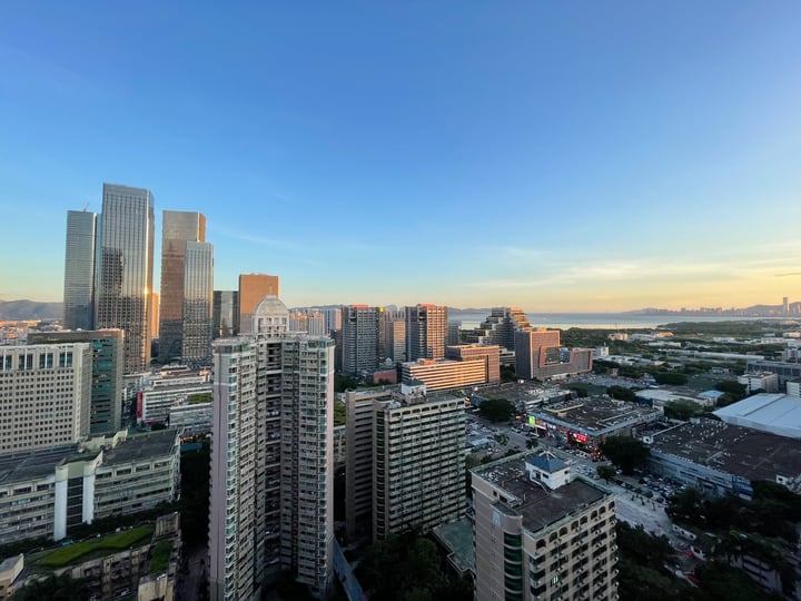 A city skyline with tall buildings, including skyscrapers, is captured during sunset, with a clear blue sky and a distant body of water.