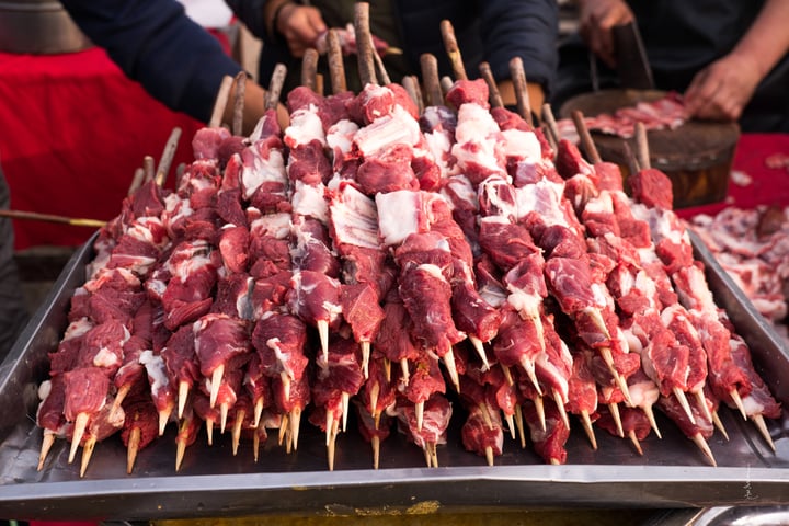 A bustling outdoor market scene features a large metal tray filled with skewered meat, including skewered beef and pork, on a table covered with a red tablecloth. The market is bustling with activity, with people in the background, and the image captures the lively atmosphere of a food market.