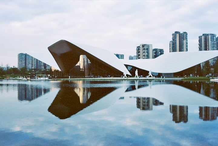 A modern building with a curved roof and white walls is reflected in a large body of water, surrounded by tall buildings and trees.