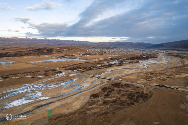An aerial view of a vast, arid landscape with a winding river, surrounded by mountains and a cloudy sky.