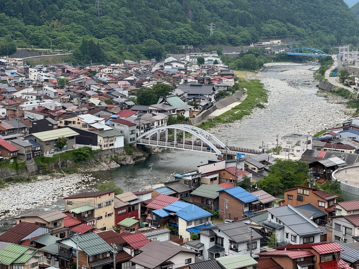 A picturesque mountain town with houses, bridges, and a river, surrounded by lush greenery and trees.