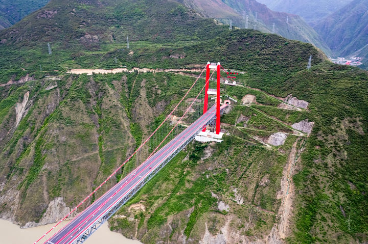 A red suspension bridge spans a river, with a red cable extending from the bridge to the river. The bridge is surrounded by green mountains and a clear blue sky.
