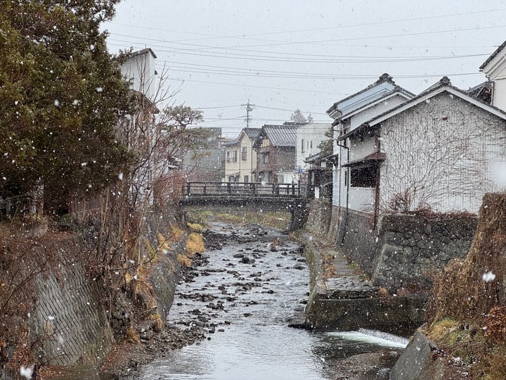 A serene winter scene in a small town features a river flowing through a stone bridge, surrounded by houses with traditional architecture. Snowflakes are falling, adding a touch of tranquility.