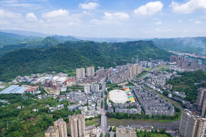 An aerial view of a city nestled between mountains, with tall buildings and a large white dome in the center, surrounded by lush greenery.