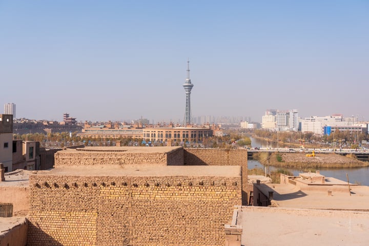The image shows a desert cityscape with a mix of traditional and modern buildings, some with solar panels and others with traditional roofs. The city is surrounded by a hazy blue sky and a distant city skyline.
