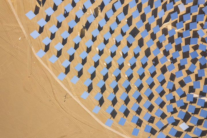 An aerial view of a large solar farm in a desert-like environment, with solar panels arranged in a circular pattern and a large white sun tower in the center. The farm is surrounded by a vast expanse of sandy terrain and a few wind turbines in the distance.