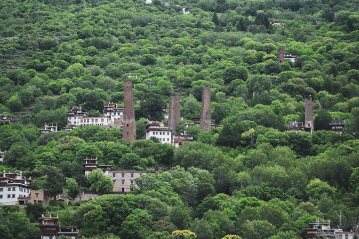 A hillside town with a mix of modern and traditional buildings, surrounded by lush greenery, is captured in a serene, aerial view.