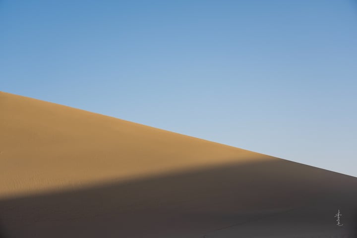 A solitary figure stands on a sandy hill in a vast desert landscape, with a clear blue sky above.