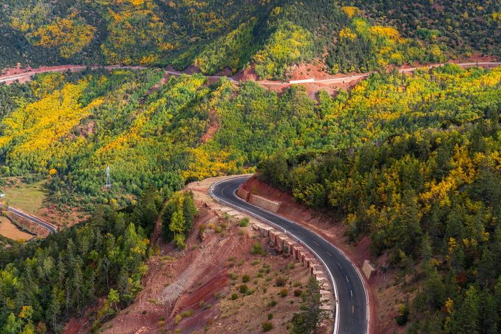 An aerial view of a winding road through a lush, green valley, surrounded by trees and hills, with a clear blue sky above.