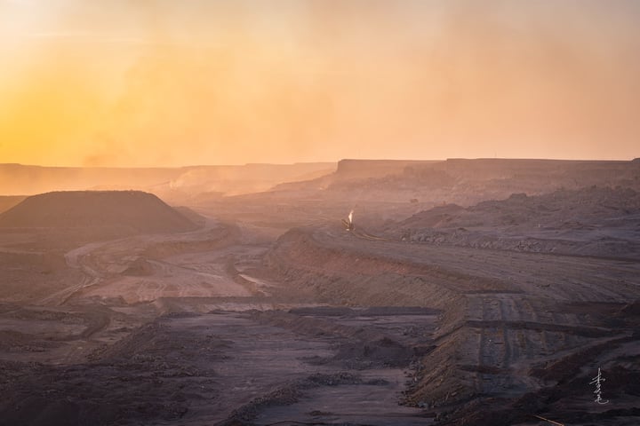 An aerial view of a desert landscape at sunset, with a dirt road winding through the landscape, a solitary flag flying in the distance, and a warm orange glow illuminating the scene.
