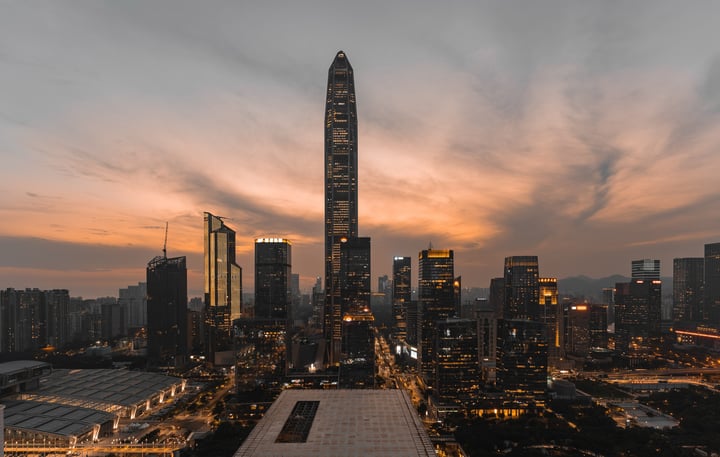 A city skyline at dusk, with a tall, modern skyscraper dominating the scene, surrounded by other tall buildings and a large, circular building. The sky is painted with orange and pink hues, and the city is illuminated by street lights.