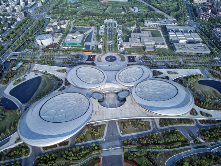 An aerial view of a modern cityscape features a large, circular building with multiple levels and a circular pool, surrounded by a network of roads and parks.