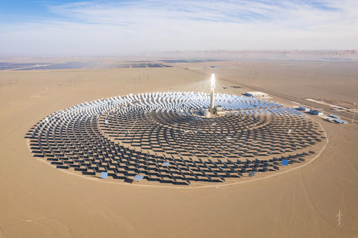 An aerial view of a large solar farm in a desert-like environment, with solar panels arranged in a circular pattern and a large white sun tower in the center. The farm is surrounded by a vast expanse of sandy terrain and a few wind turbines in the distance.