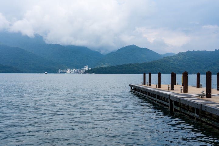 A wooden pier extends into a calm lake, with a cityscape in the distance and mountains covered in greenery.