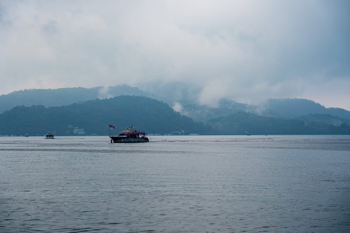 A serene scene of a large body of water with a small boat, a red and white flag, and a mountain range in the background.