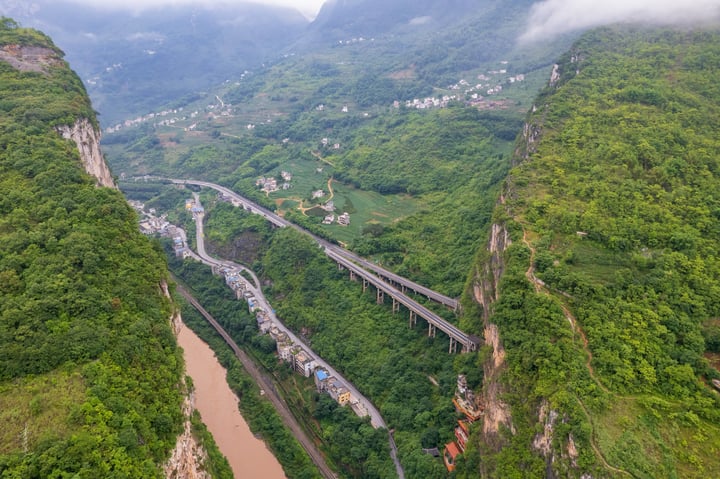 An aerial view of a mountainous valley with a winding river, a bridge, and a town in the distance.