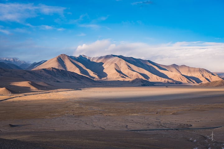 A vast desert landscape with sand dunes and mountains, a clear blue sky with clouds, and a few distant trees.